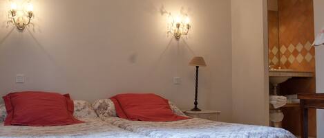 Quarto com uma cama grande, duas almofadas vermelhas, um candelabro e uma mesa de cabeceira com abajur.