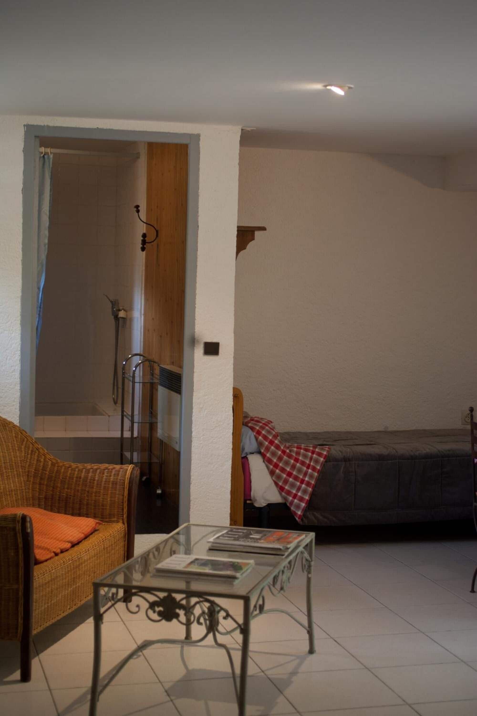 Een kamer met een rieten bank, een glazen salontafel, een bed met een rood geruit plaid en een douchegedeelte met een houten deur.