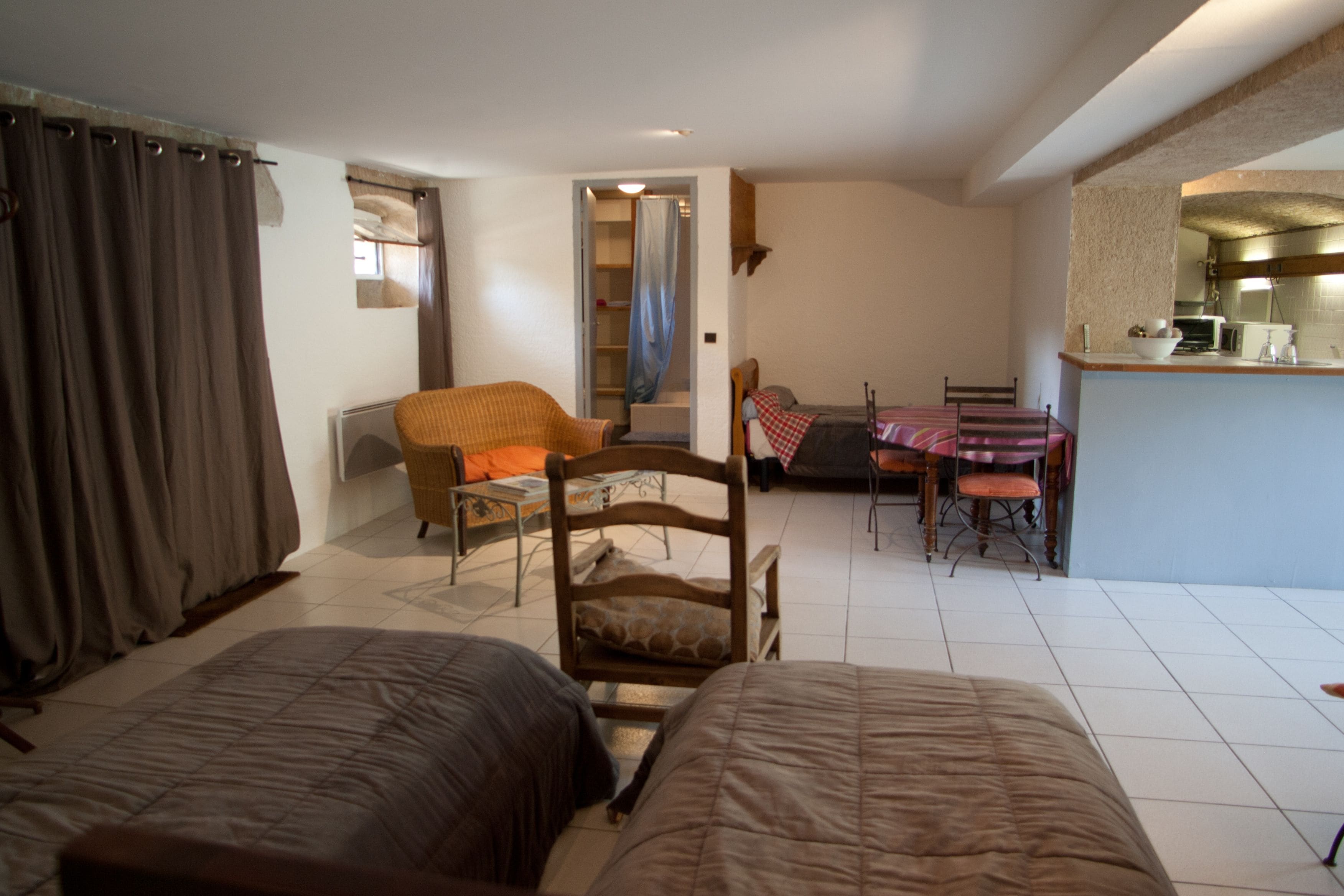A bedroom with two beds, a chair, a dining table with chairs, and a kitchen area.