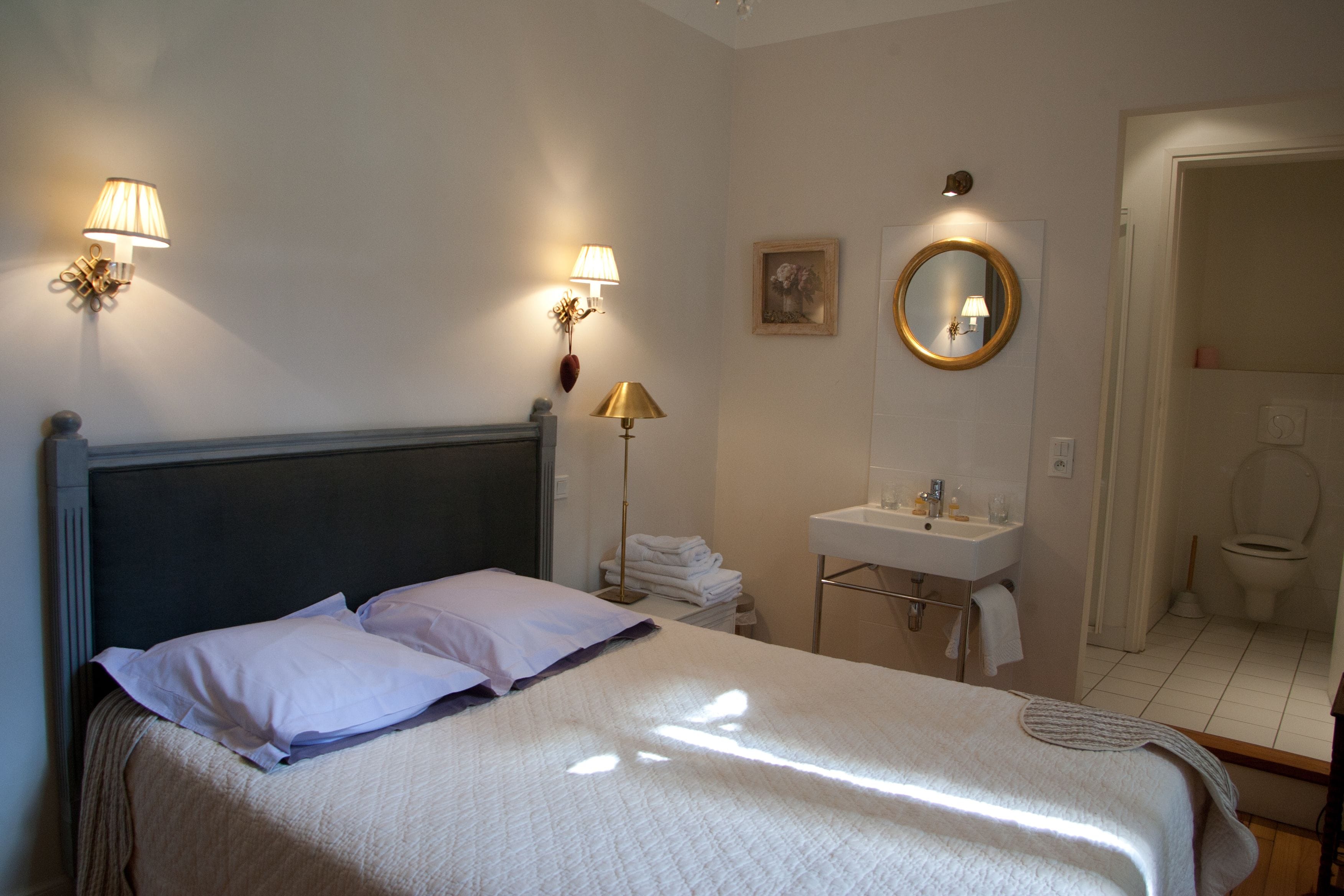 Une chambre d’hôtel comprenant un lit, une lampe de chevet, un miroir fixé au mur et une salle de bain avec un lavabo et des toilettes.