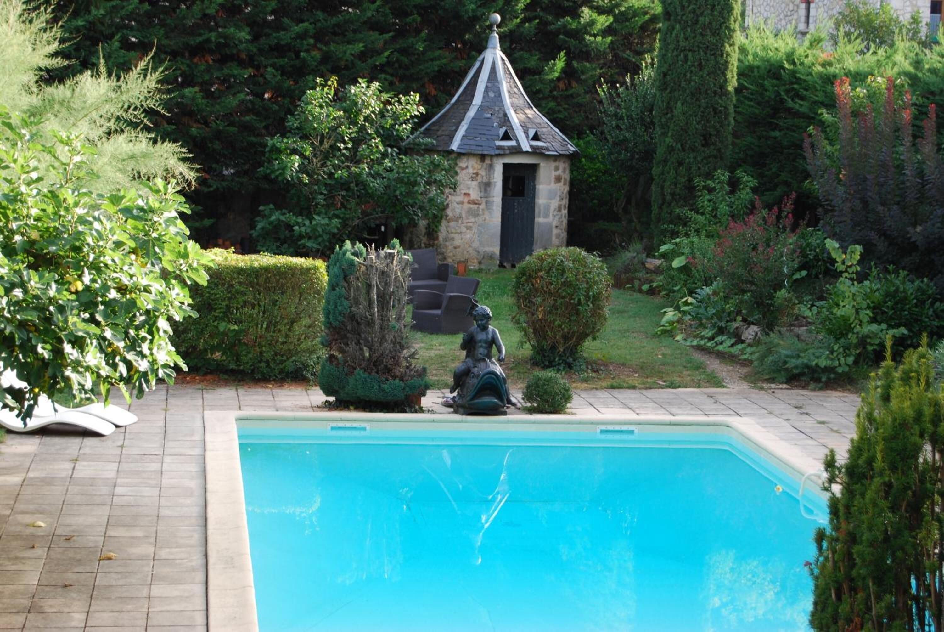 Une piscine ornée d’une statue au centre, entourée de haies bien entretenues et, au fond, d’un bâtiment en pierre doté d’un toit en forme de flèche.