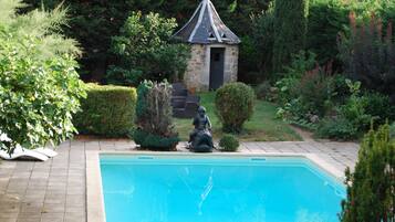 Une piscine ornée d’une statue au centre, entourée de haies bien entretenues et, au fond, d’un bâtiment en pierre doté d’un toit en forme de flèche.