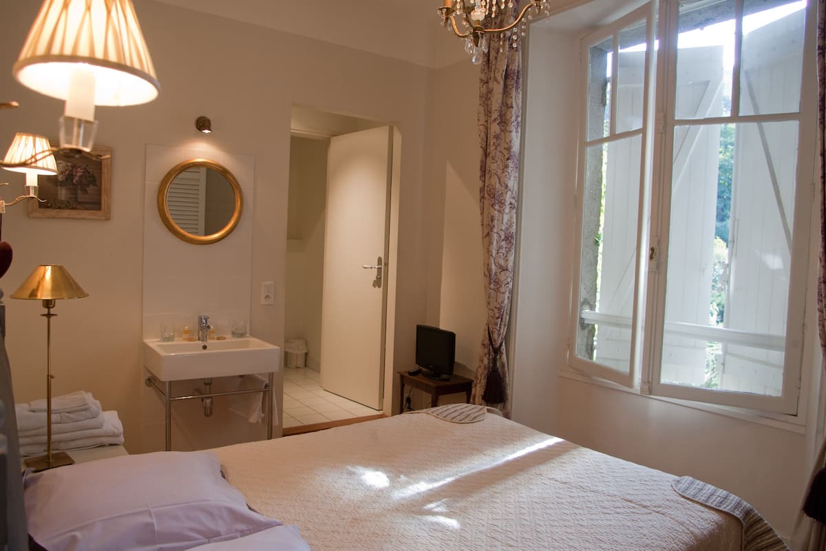 Une chambre d’hôtel comprenant un lit, une table de chevet avec une lampe, un miroir, une fenêtre avec des rideaux et une salle de bain avec un lavabo et un porte-serviettes.