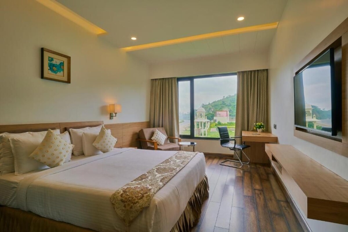 A hotel room with a large bed, a desk, a chair, a TV, and a window with a view of greenery.