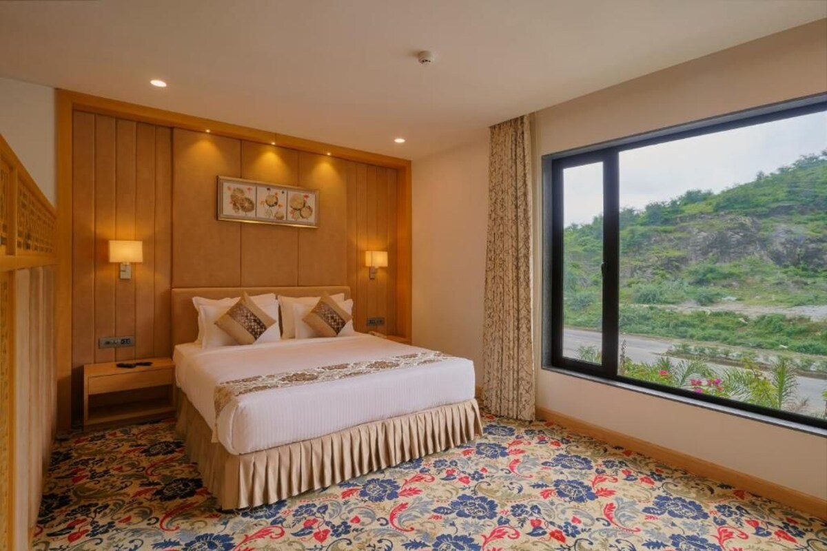 A hotel room with a large bed, wooden paneling, a view of greenery, and a patterned carpet.
