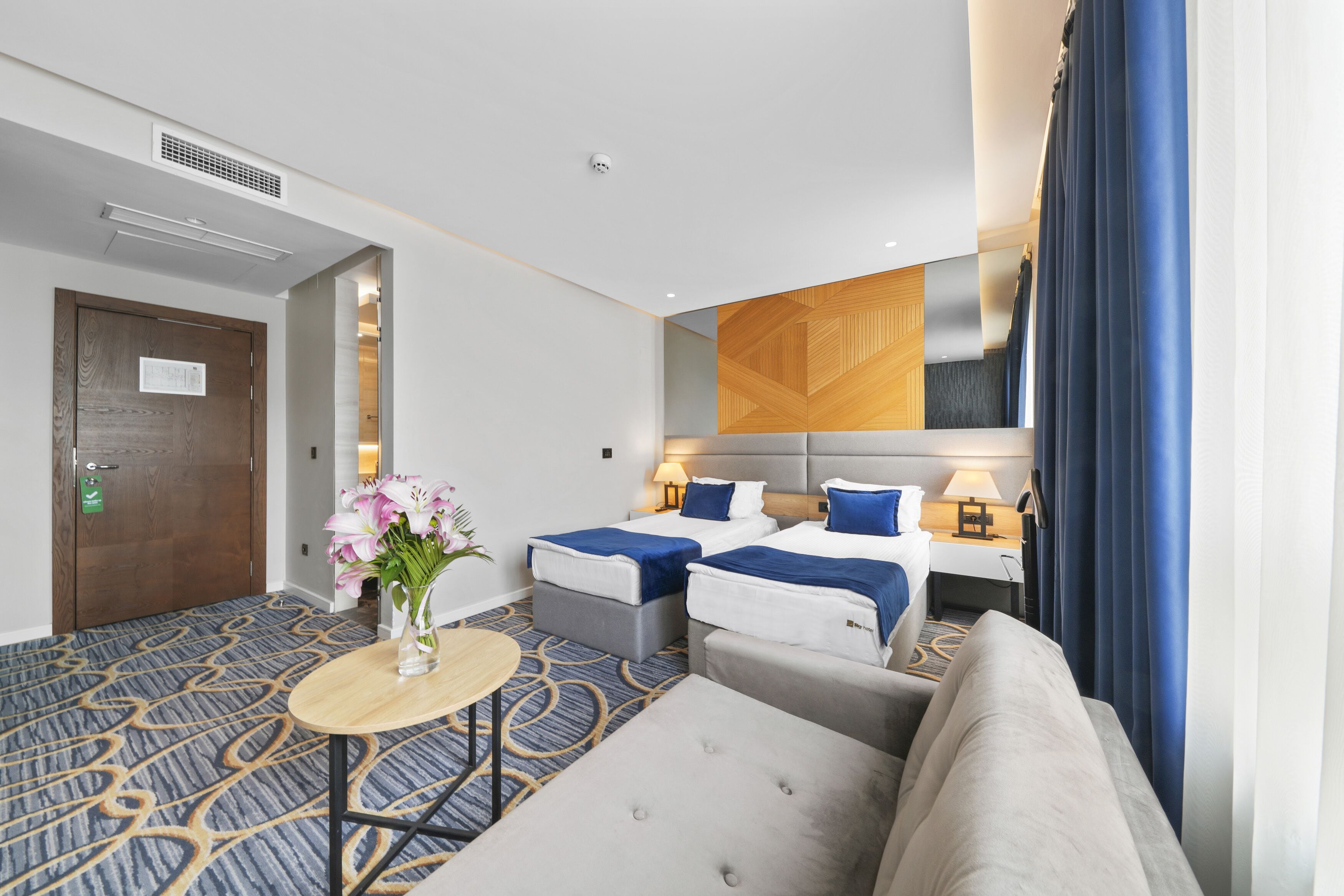 Hotelkamer met twee bedden, een bank, een kleine tafel met een bloemetjesvaas en een grote spiegel.