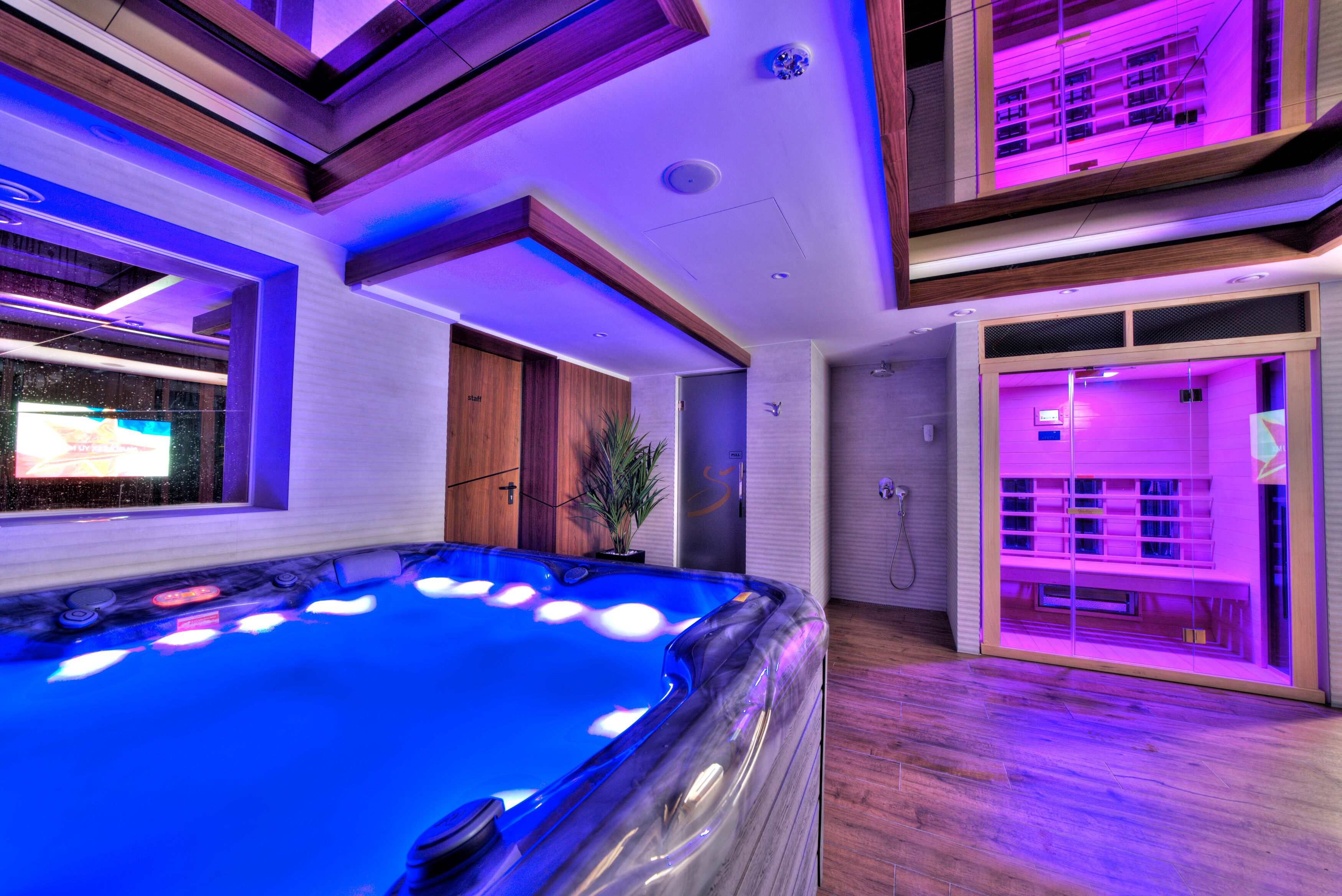 A modern indoor swimming pool with a wooden floor, surrounded by a multi-level room with wooden beams and purple lighting.
