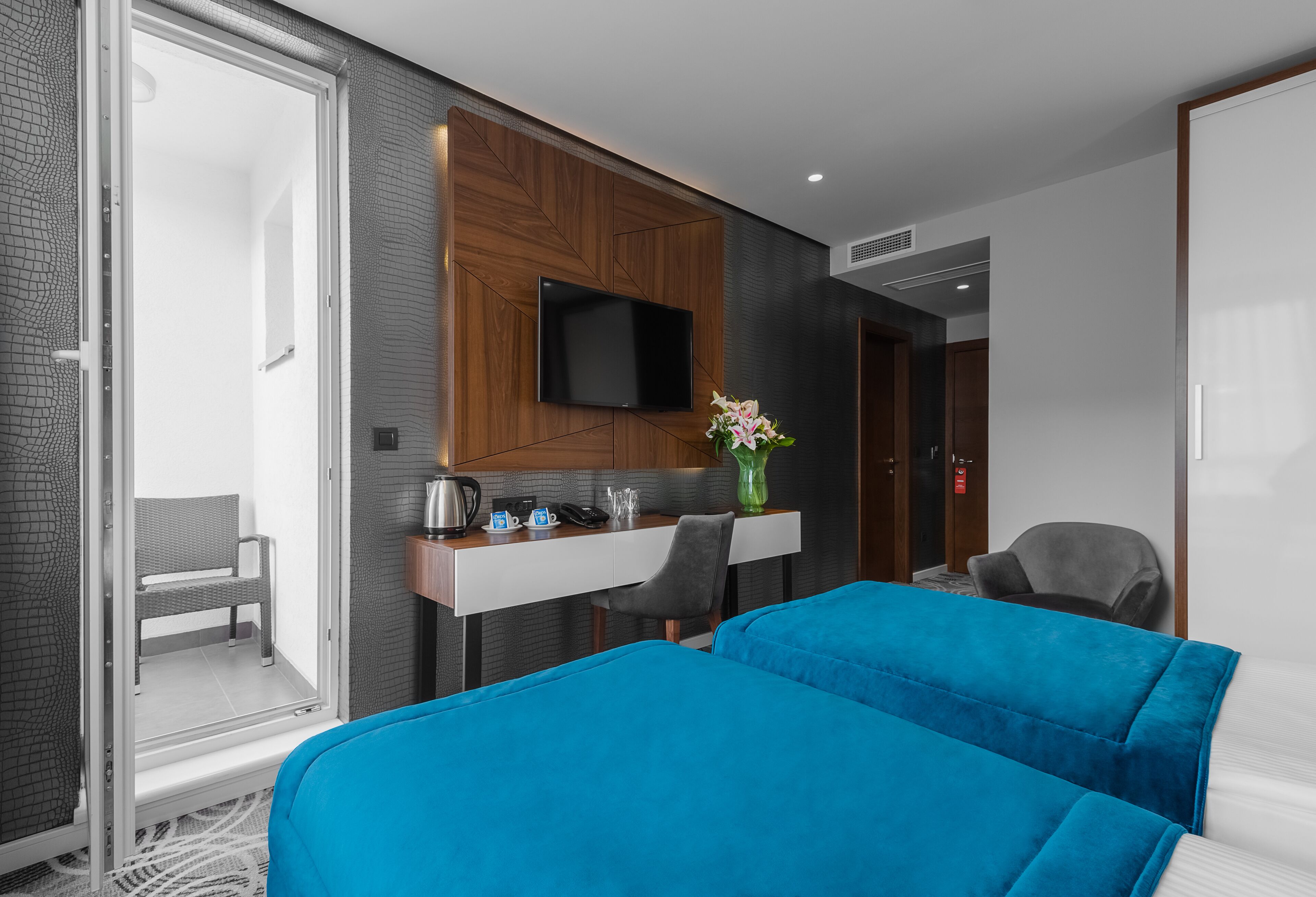 A modern hotel room with a large bed, a desk with a flat-screen TV, a chair, and a small seating area with a bench.