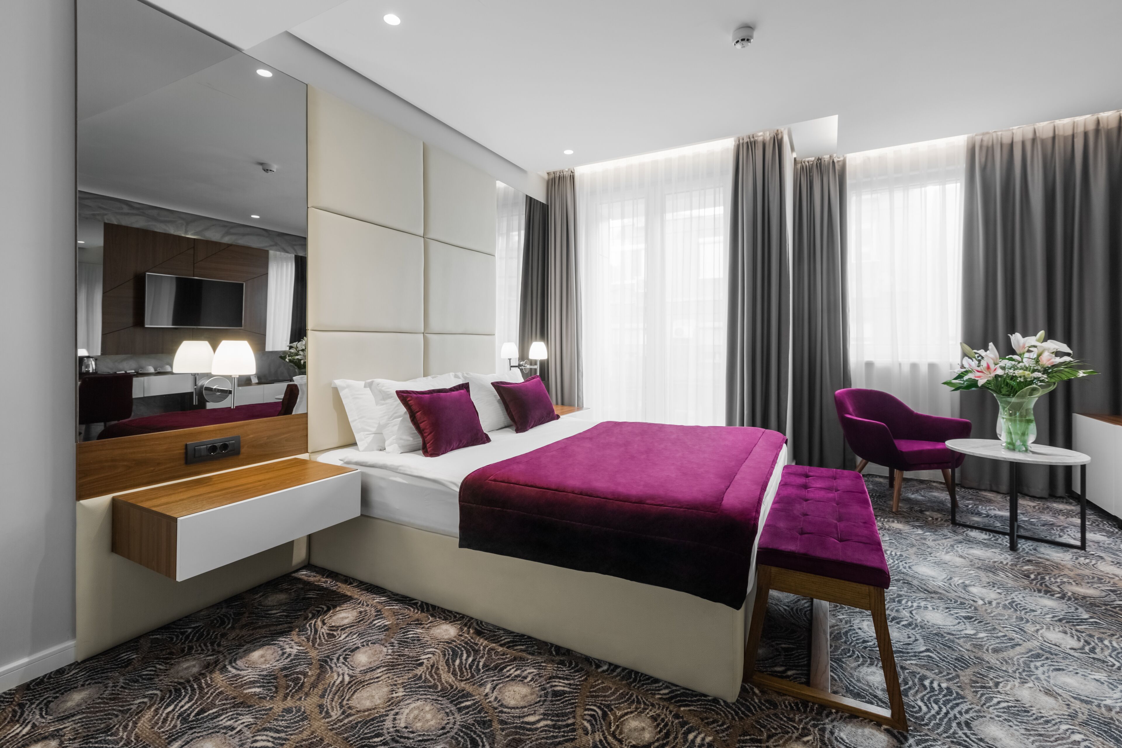 Una habitación de hotel moderna con una cama grande, una mesita de noche, un televisor y una zona de estar con una silla y una pequeña mesa.