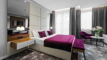 Una habitación de hotel moderna con una cama grande, una mesita de noche, un televisor y una zona de estar con una silla y una pequeña mesa.