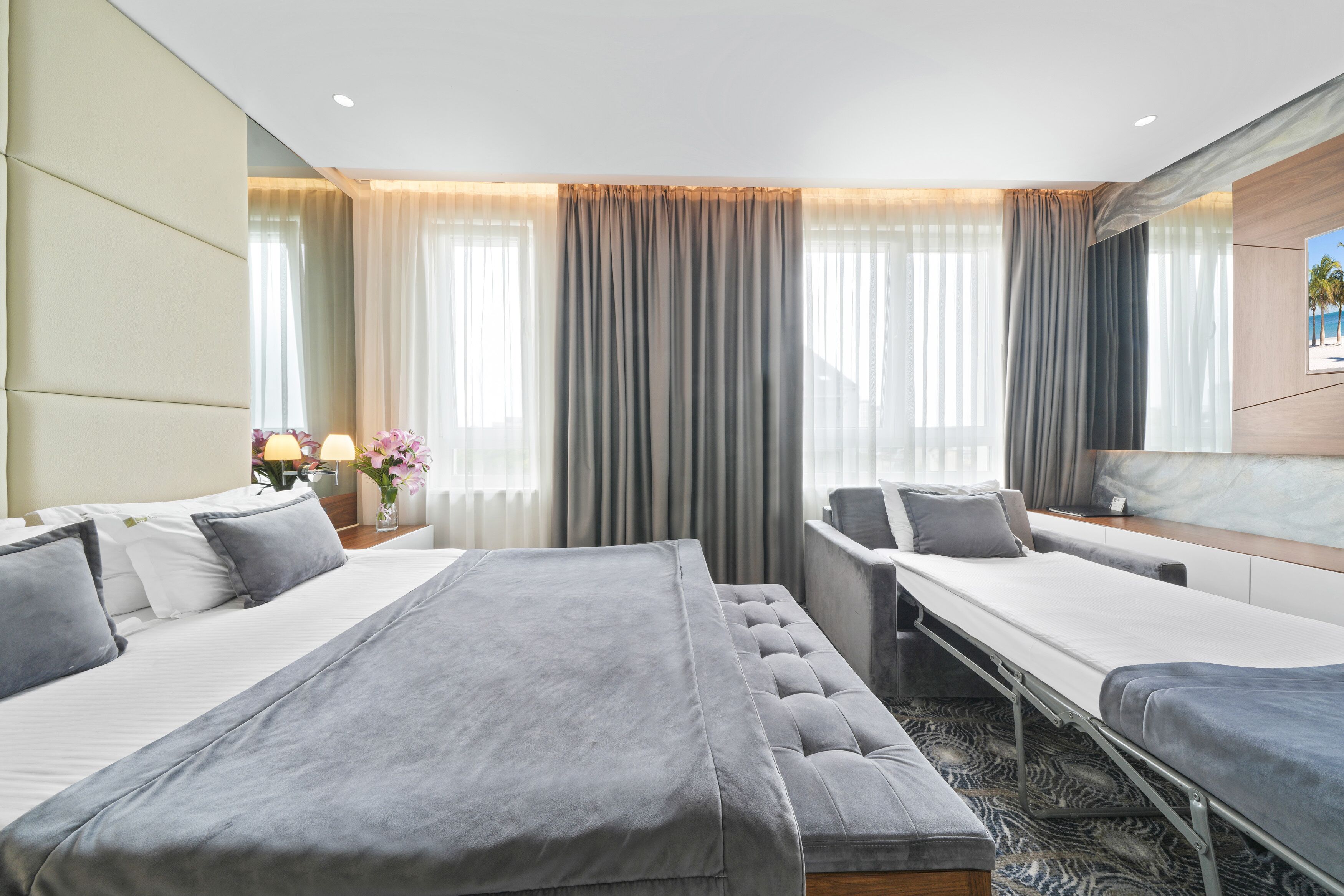 A modern hotel room with two beds, a sofa, a desk, and a large window with curtains.