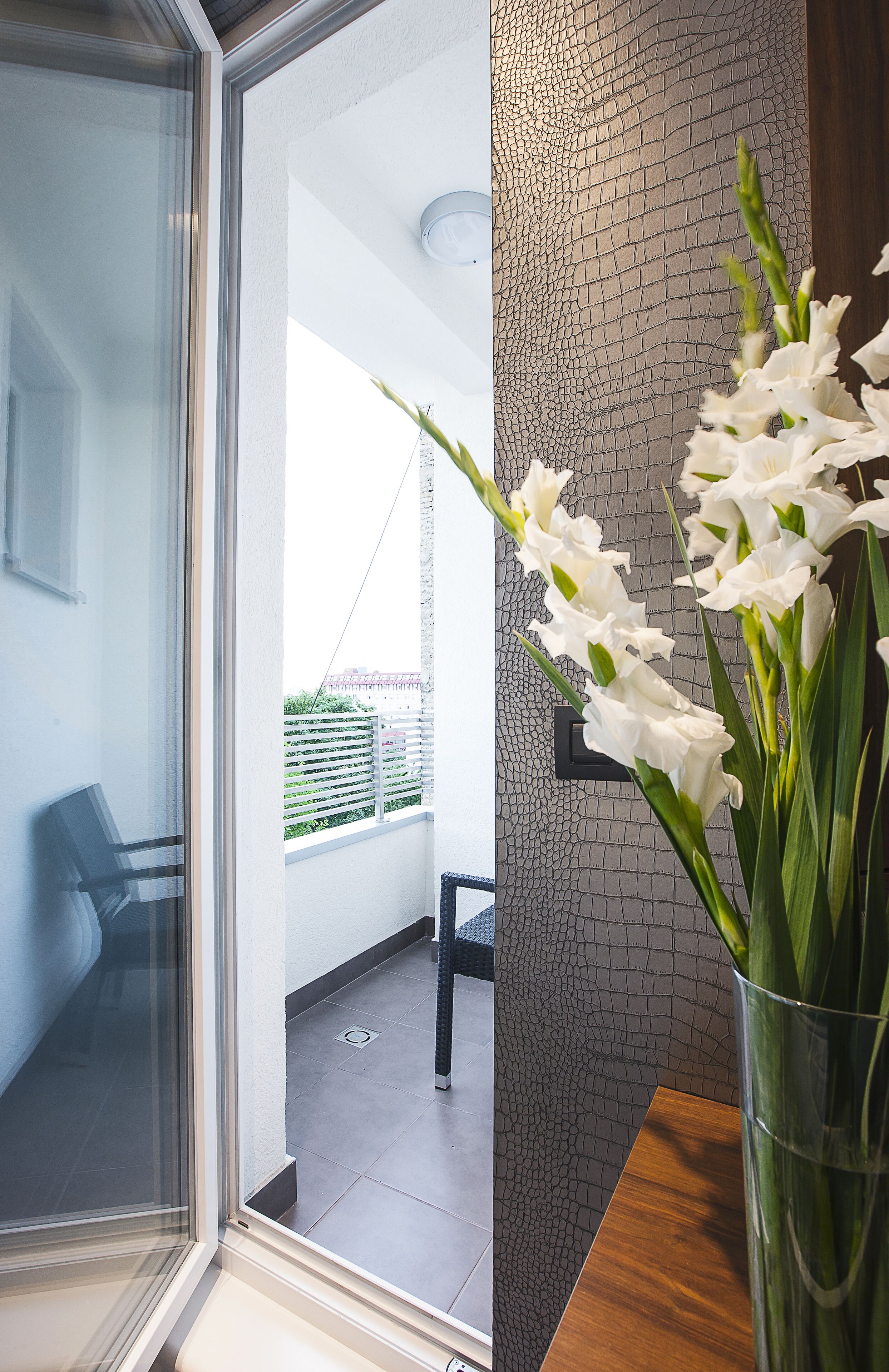 A modern interior with a glass door leading to a balcony, a textured wall, and a vase with white flowers.