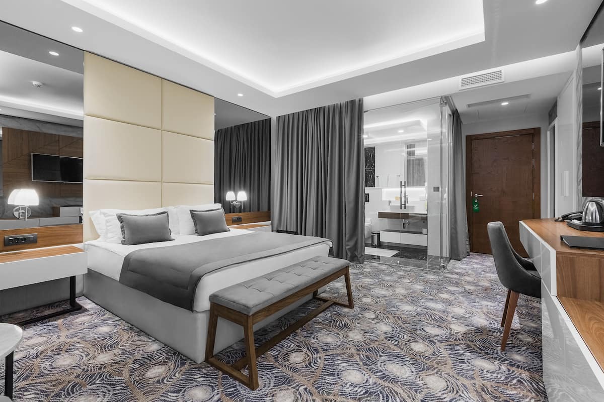 A modern hotel room with a large bed, bedside tables, a bench, a desk, and a mirror.