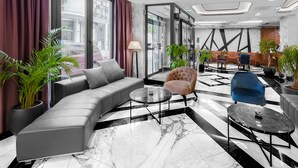 A modern hotel lobby with a grey sectional sofa, a blue armchair, and a black coffee table. There are potted plants and a large window with curtains. - Sky hotel (Belgrade)