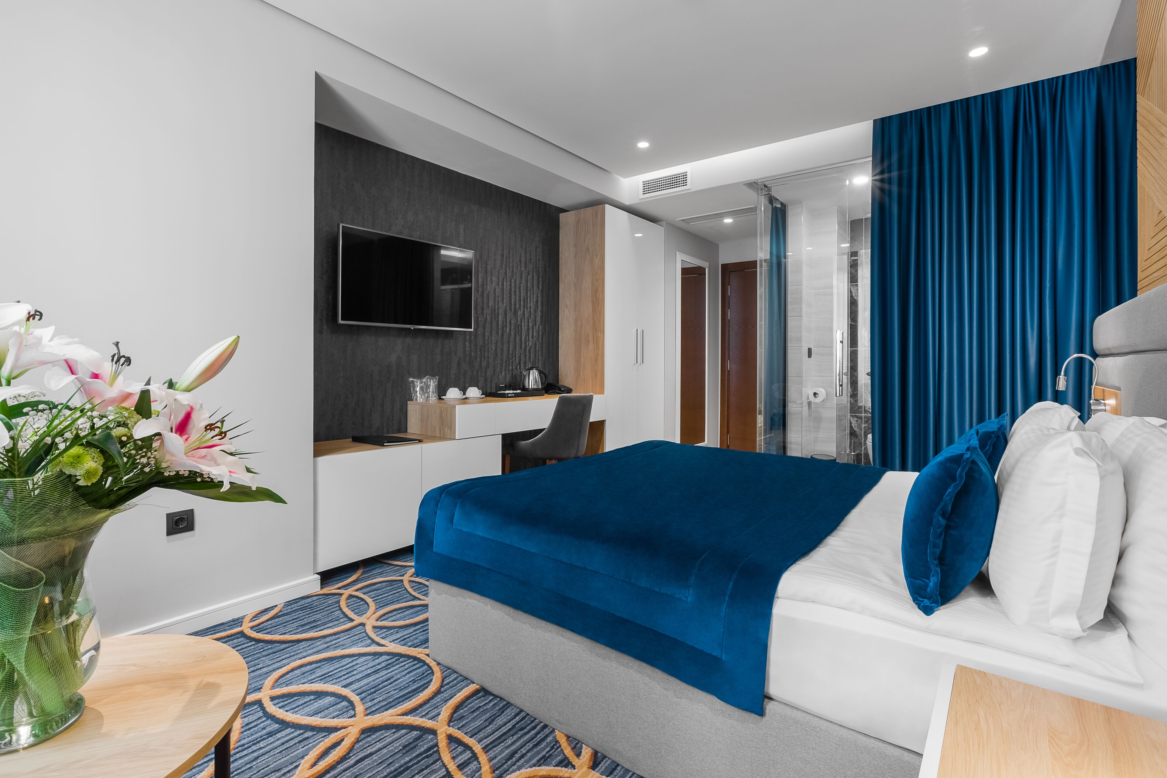 Een moderne hotelkamer met een bed, een bureau, een televisie en een badkamer, zichtbaar door een glazen deur.