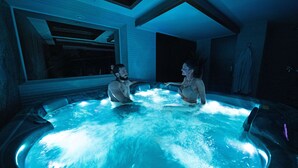 A hot tub with two people sitting inside, surrounded by a modern interior with a large window and a towel rack. - Sky hotel (Belgrade)