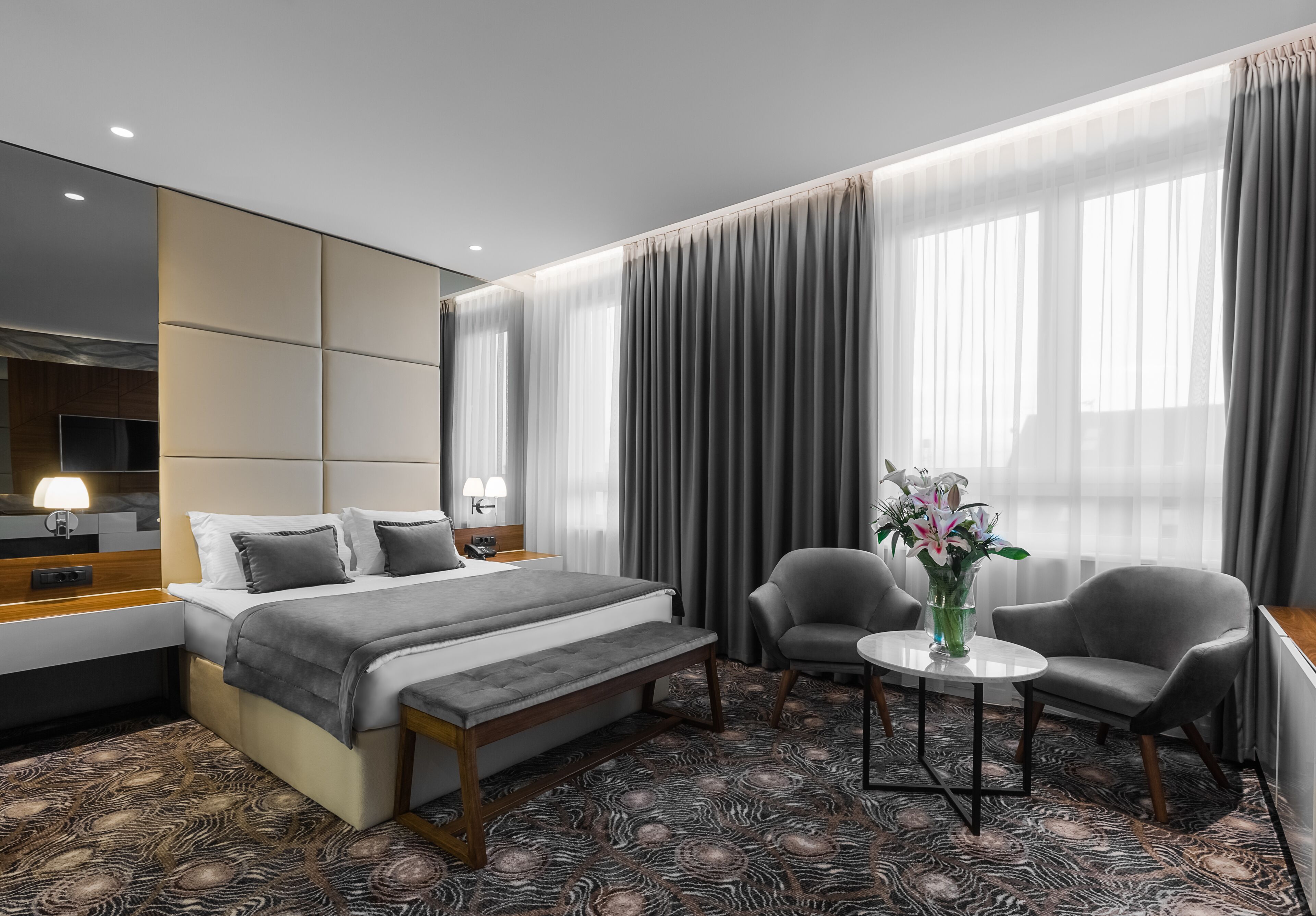 Habitació d'hotel moderna amb un llit gran, dues butaques, una taula petita i una finestra ampla amb cortines.