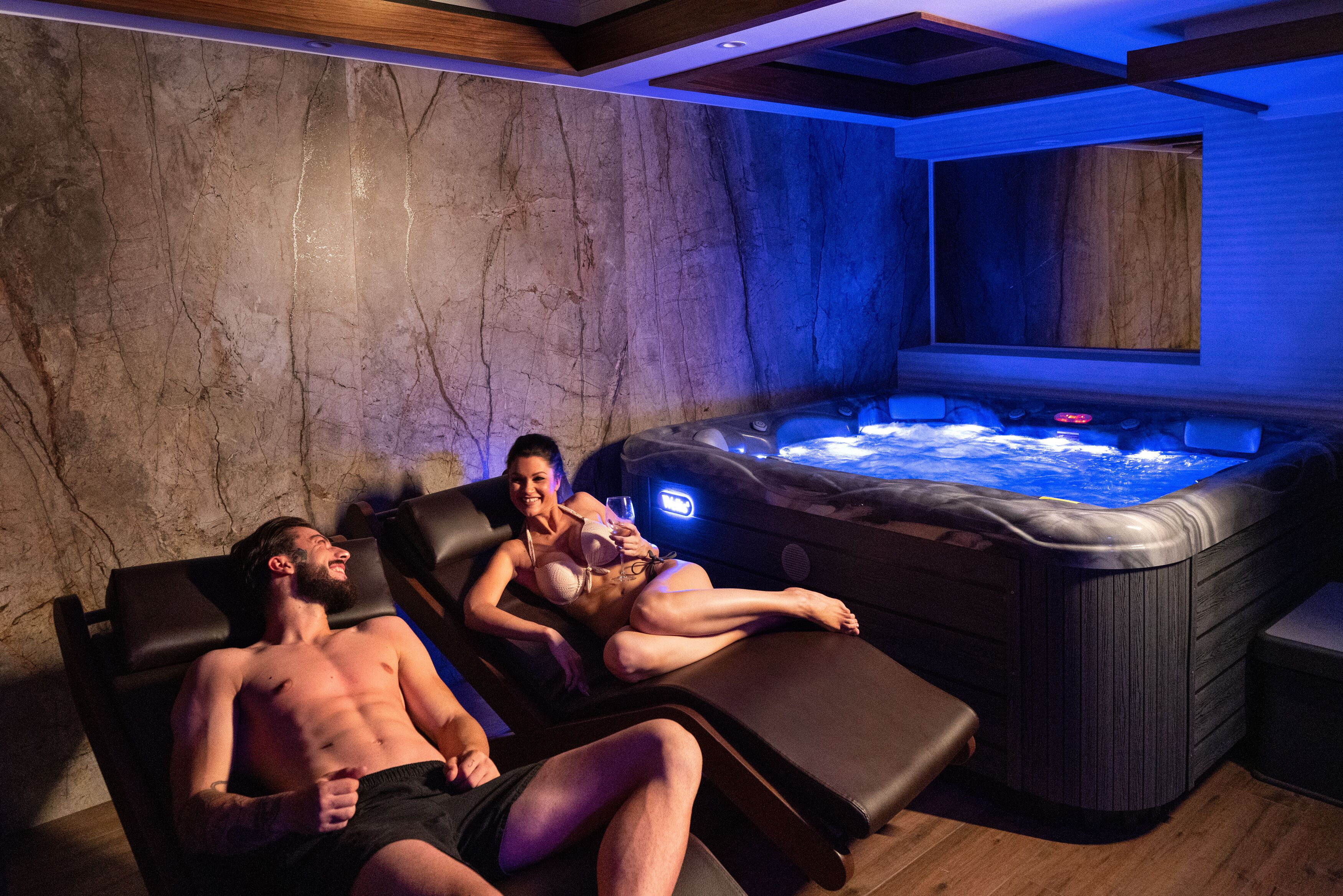 A spa area with two reclining chairs, a hot tub, and a stone wall.