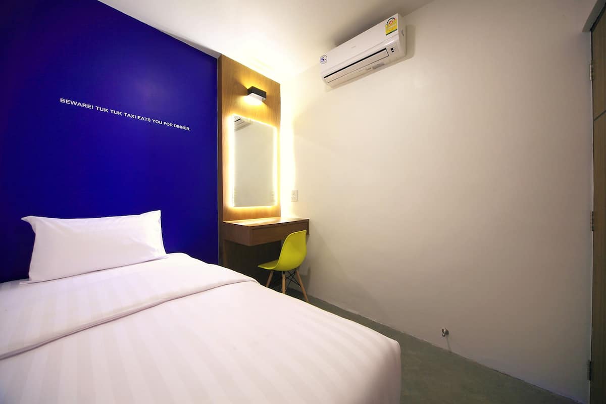 A hotel room with a bed, a desk with a chair, a mirror, and an air conditioning unit.