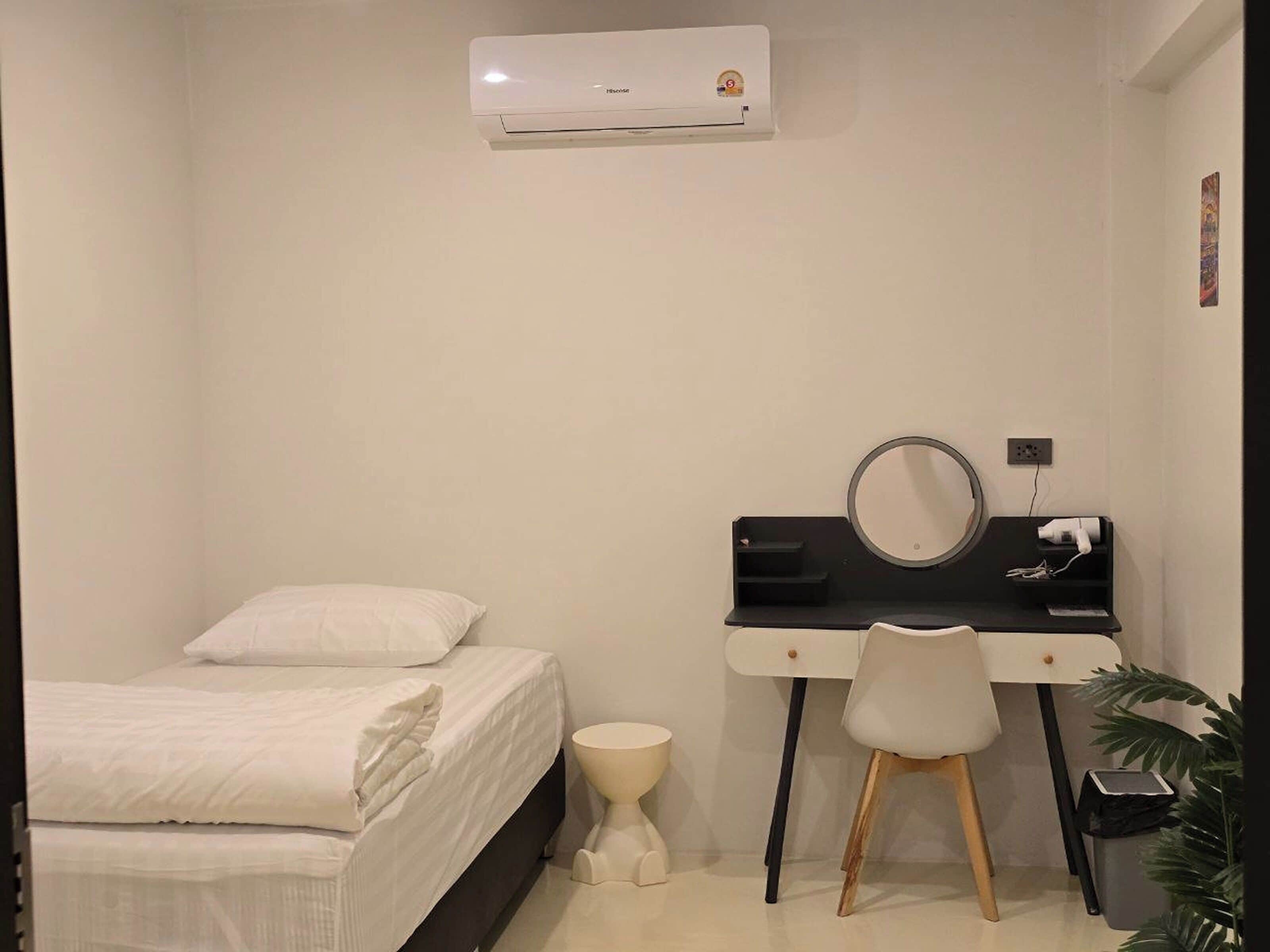 A small, clean room with a bed, a desk with a chair, a toilet, and an air conditioner.