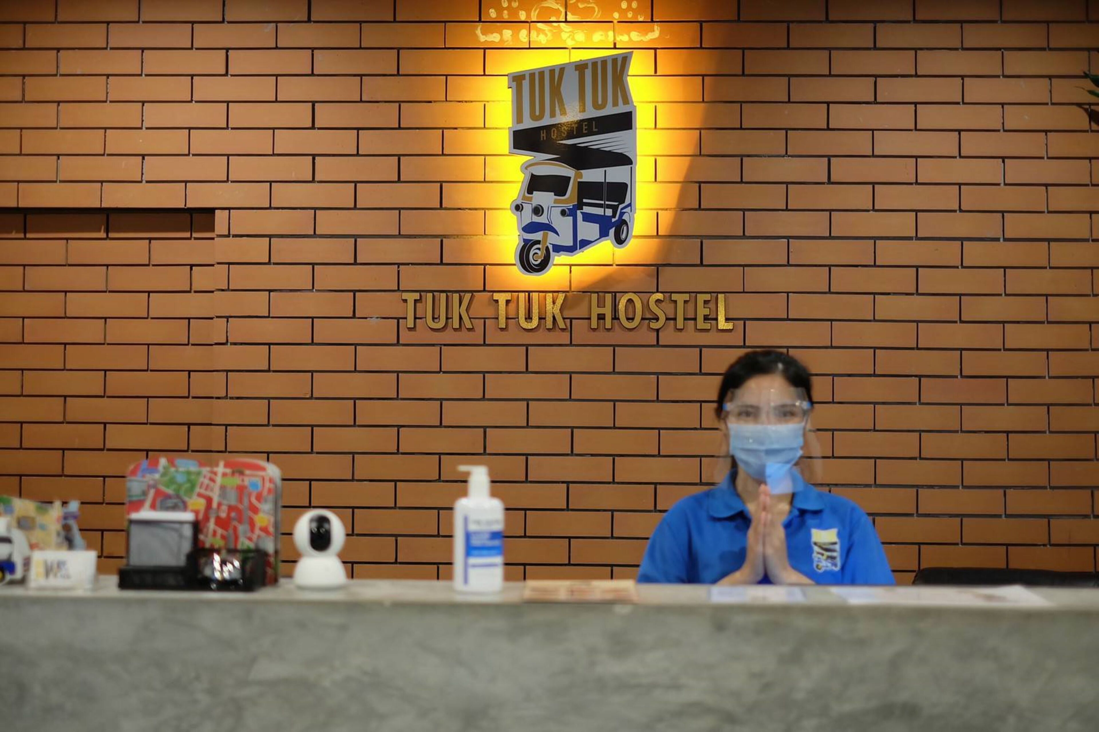 A brick wall with a sign reading "TUK TUK HOSTEL" and a person in a blue uniform standing behind a counter with a sanitizer bottle and a small TV.