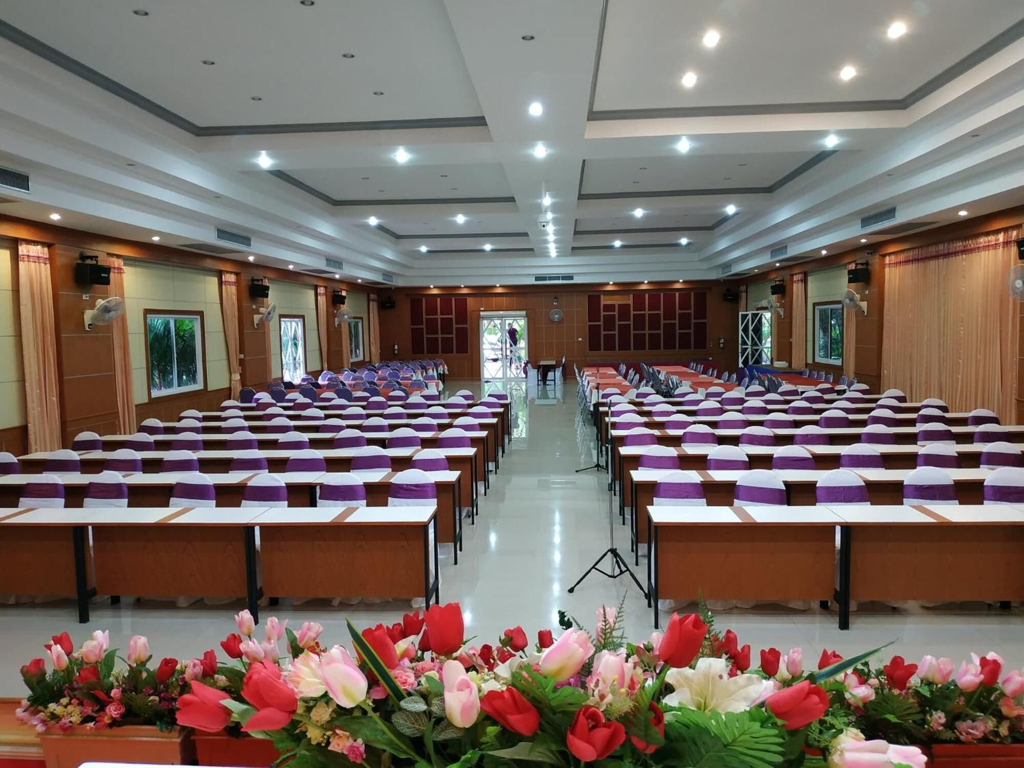A large conference room with rows of tables and chairs, a stage at the front, and a floral arrangement at the front.