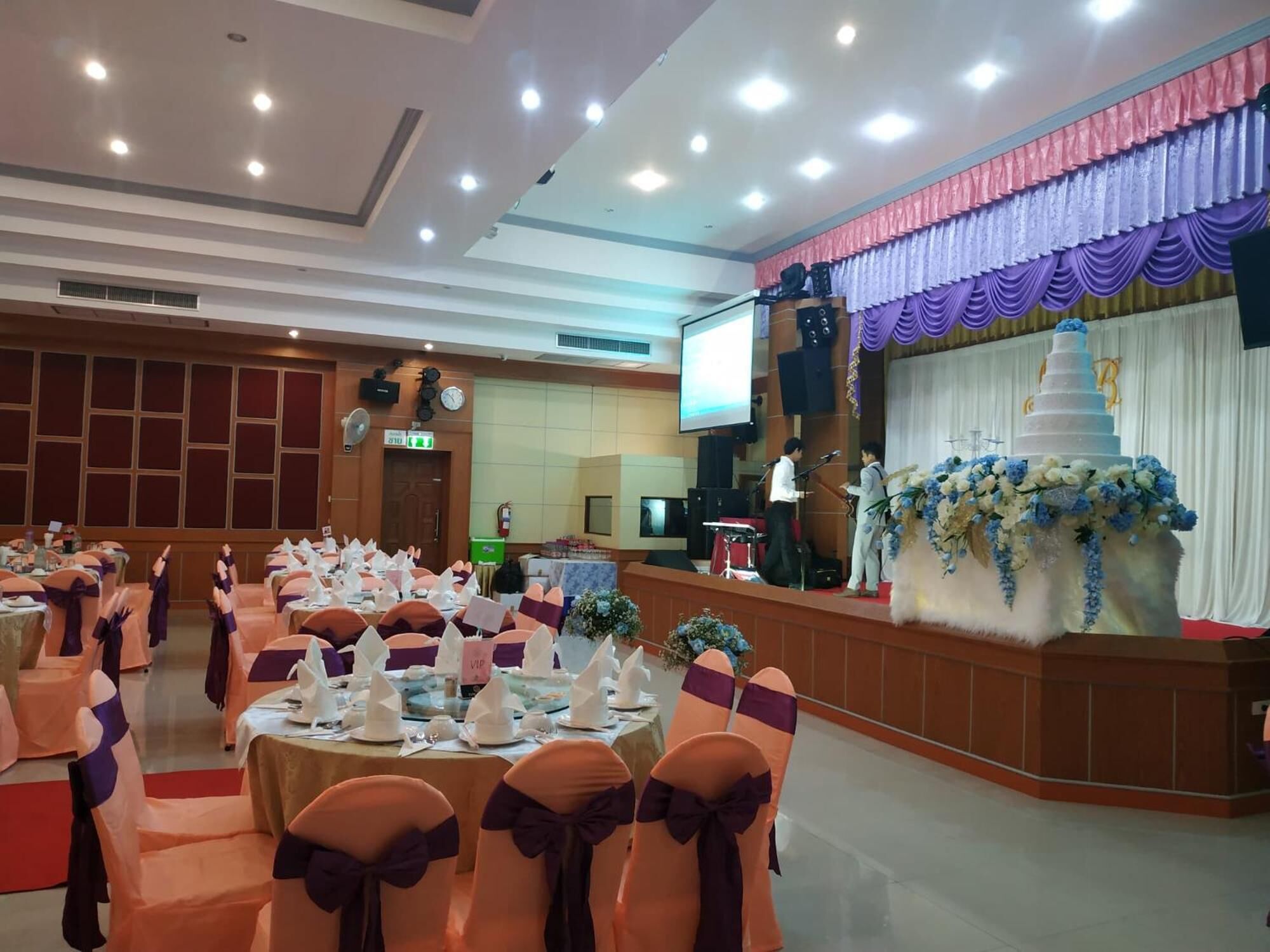 Un salón de banquetes con mesas redondas, sillas adornadas con cintas moradas, un escenario con un pastel de varios niveles y una pantalla grande.