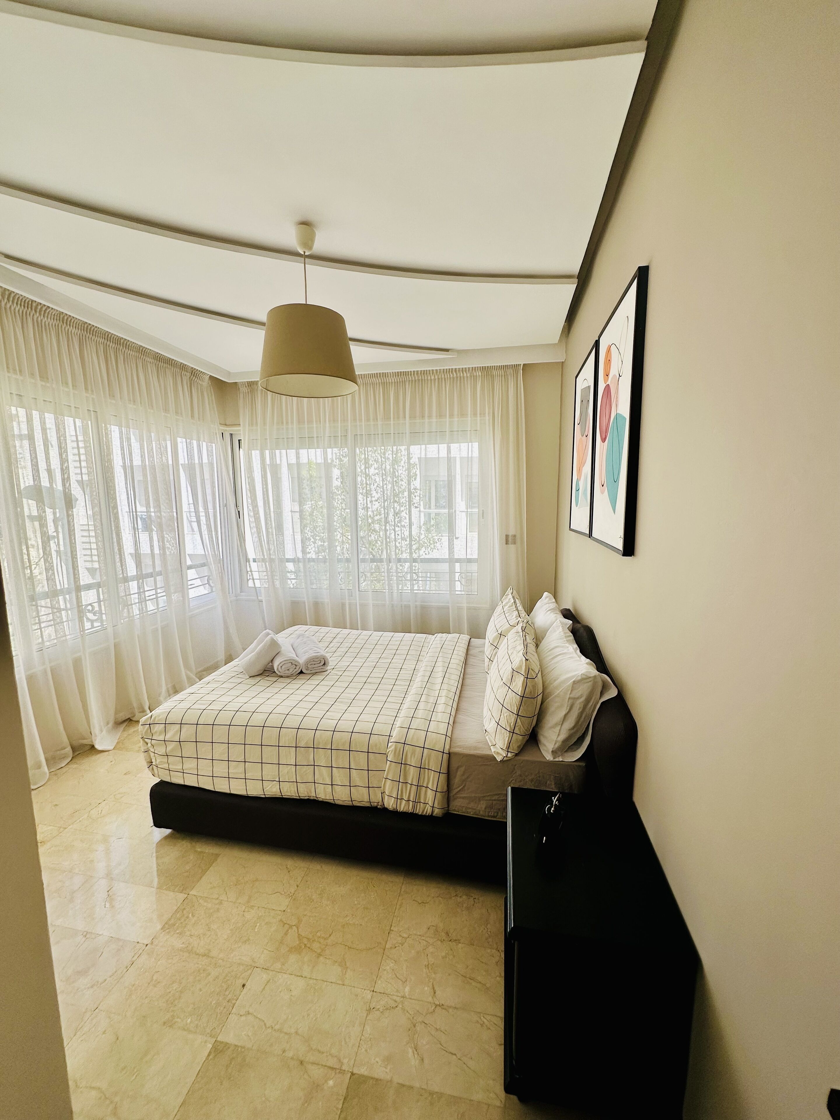 A bedroom with a bed, a ceiling light, a window with curtains, and a wall with framed pictures.