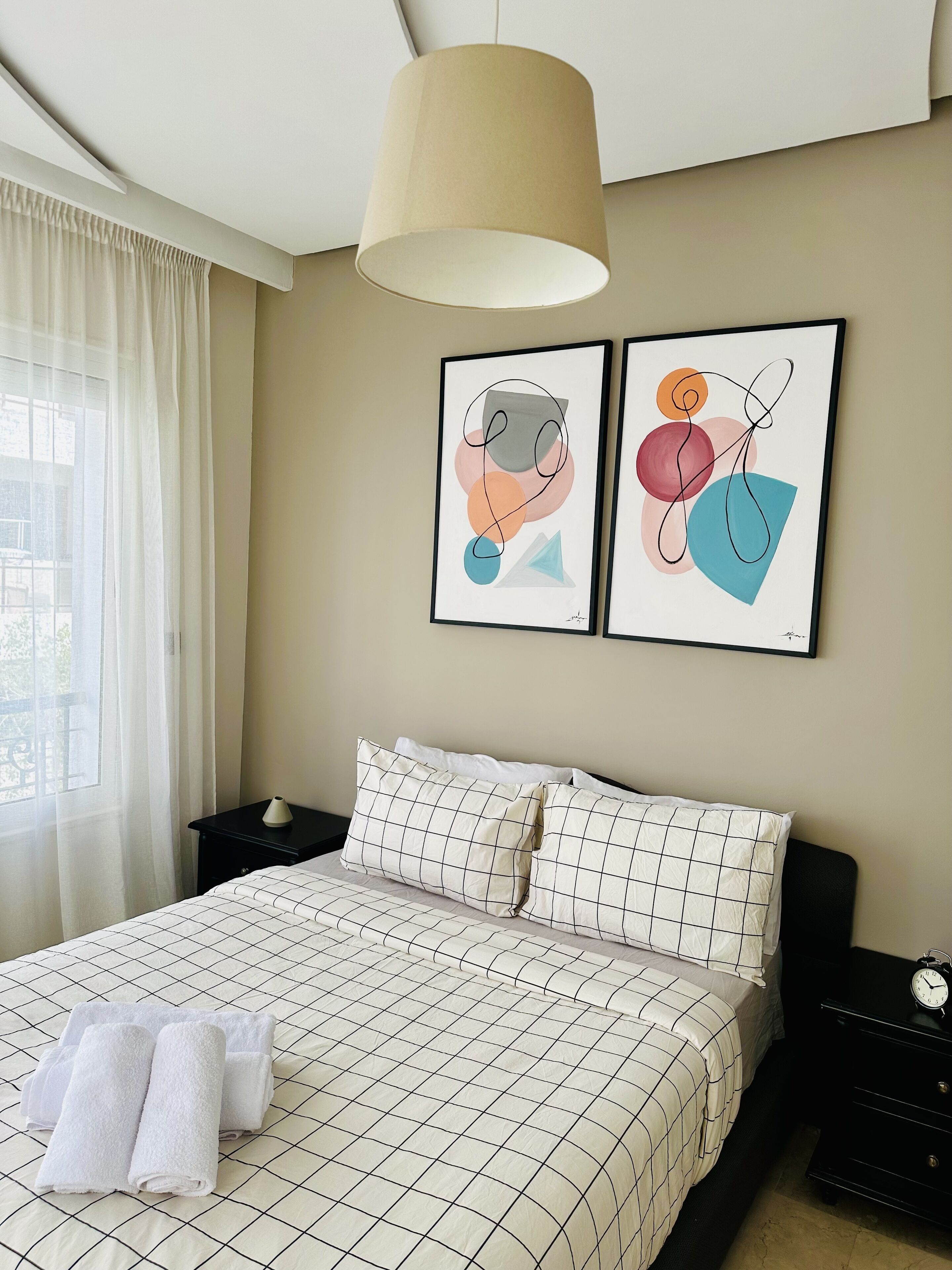 A neatly made bed with a checkered bedspread, two pillows, and rolled towels on a nightstand. There are two framed abstract paintings on the wall above the bed.