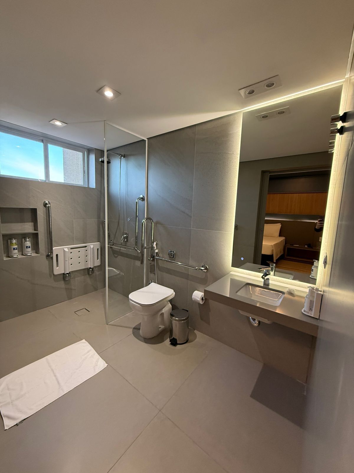 A modern bathroom with a glass-enclosed shower, a toilet, a sink with a mirror, and a towel on the floor.