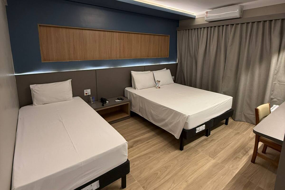 A hotel room with two single beds, a desk, a chair, a nightstand, and a wall-mounted air conditioner.