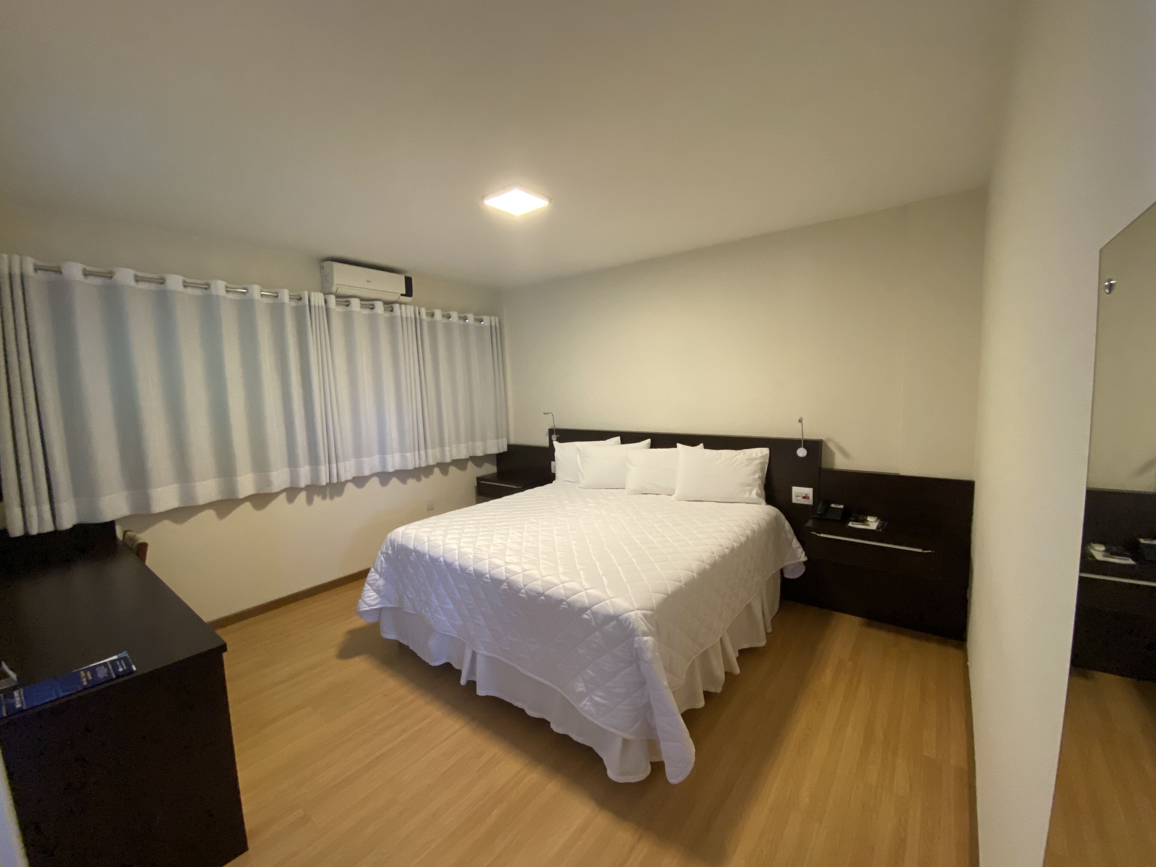 A hotel room with a large bed, a bedside table, a nightstand, a mirror, and a window with curtains.