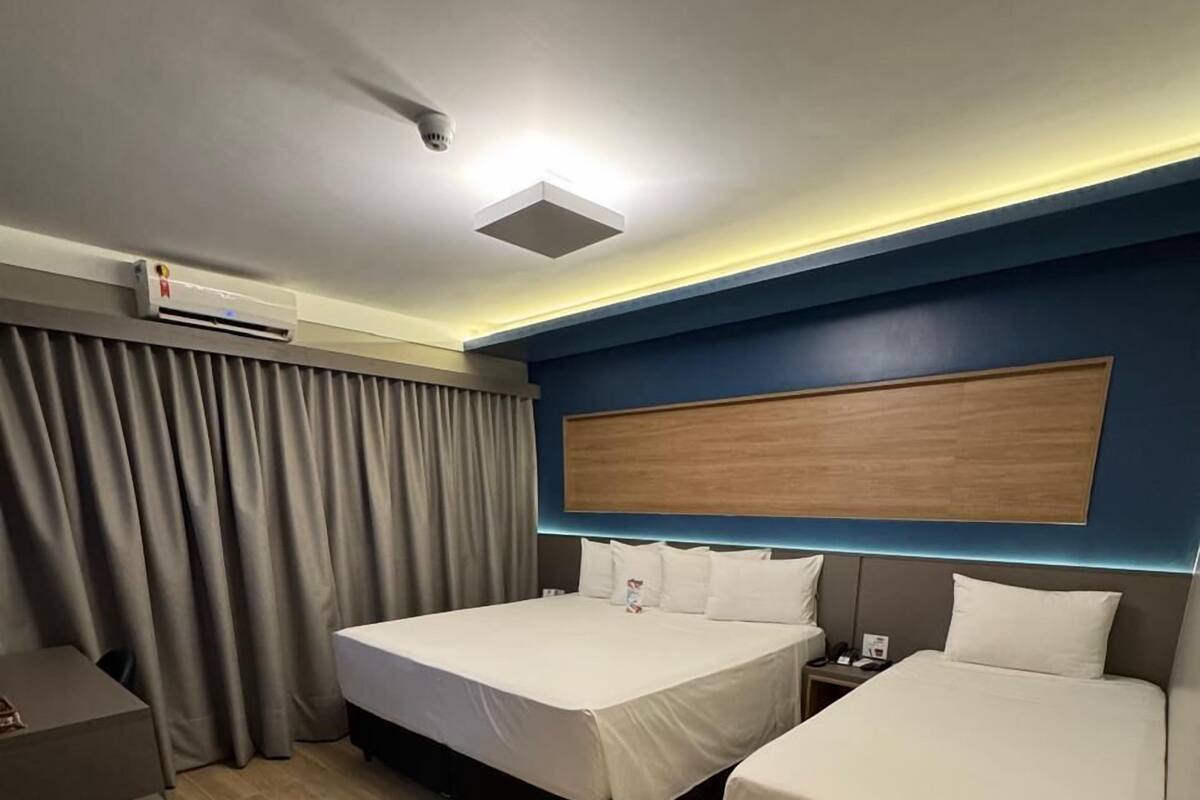 A hotel room with two beds, a desk, an air conditioning unit, and a curtain.