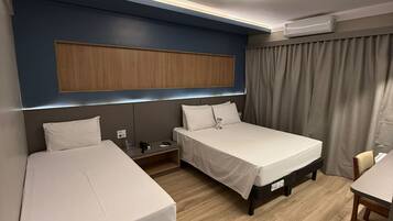 A hotel room with two single beds, a wooden headboard, a bedside table with a lamp, and a window with curtains.