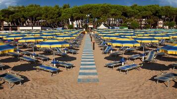 A beach with rows of sun loungers and umbrellas, a central pathway, and trees in the background.