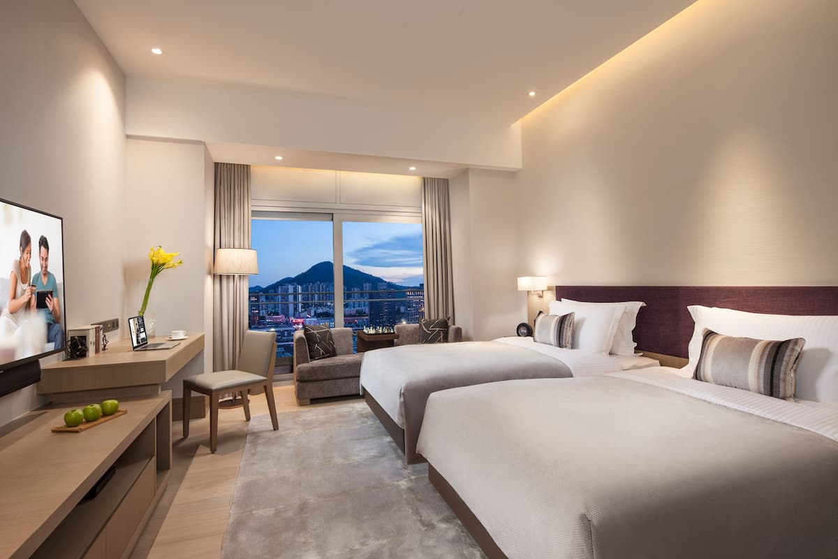 A modern hotel room with two beds, a desk with a TV, a sofa, and a view of a cityscape.