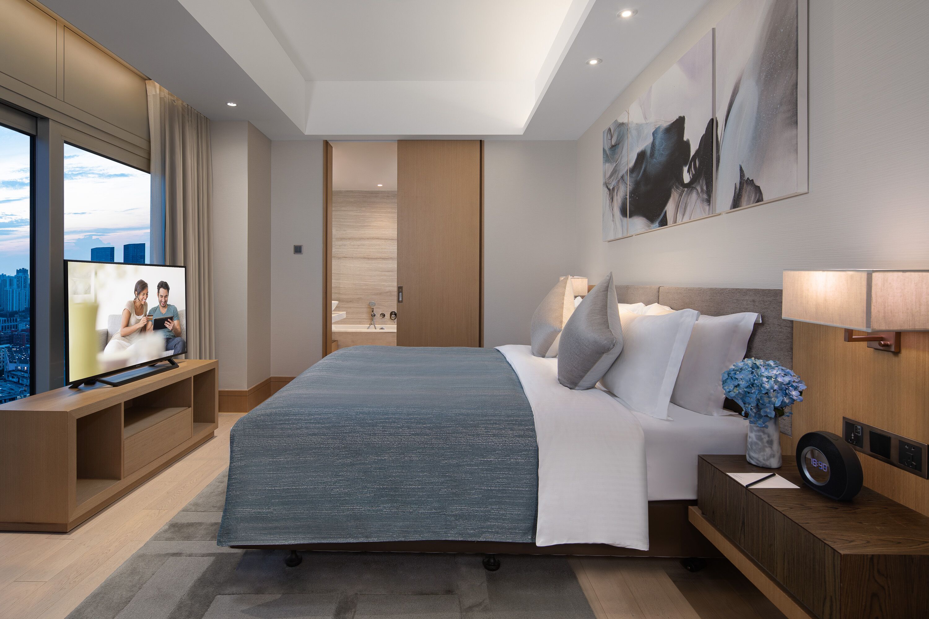 A modern hotel room with a large bed, a TV on a wooden console, a nightstand with a clock, and a city view through large windows.