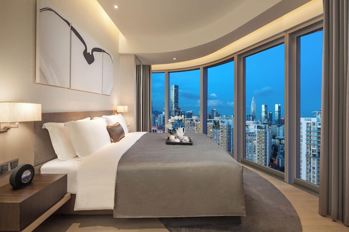 A modern hotel room with a large bed, a city view, and a digital clock on the nightstand.