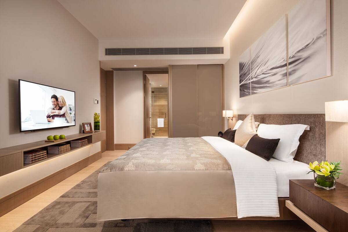 A modern hotel bedroom with a large bed, a flat-screen TV, a bedside table with a lamp, and a large abstract painting on the wall.