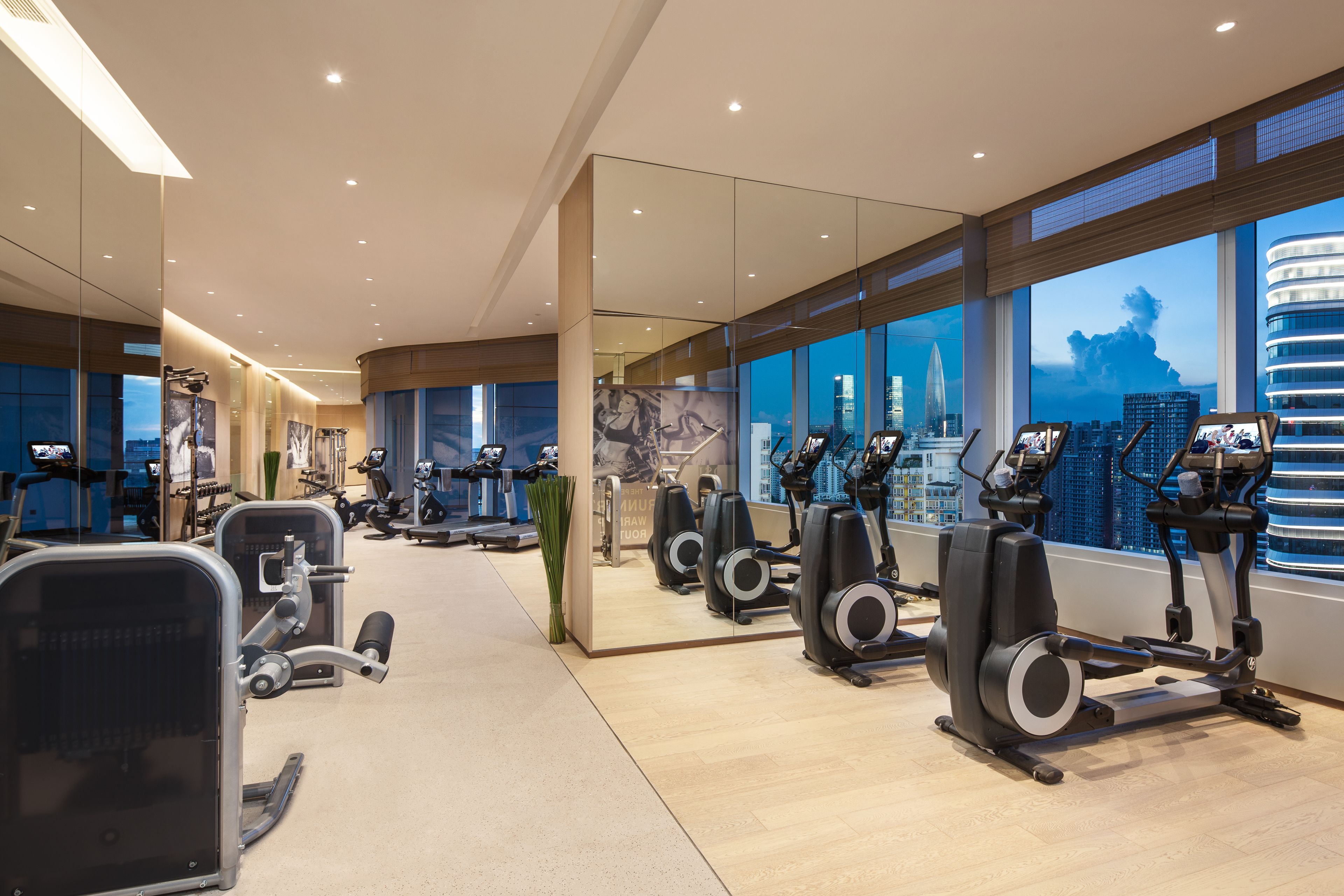 A modern gym with various exercise equipment, large windows with a city view, and a clean, spacious layout.