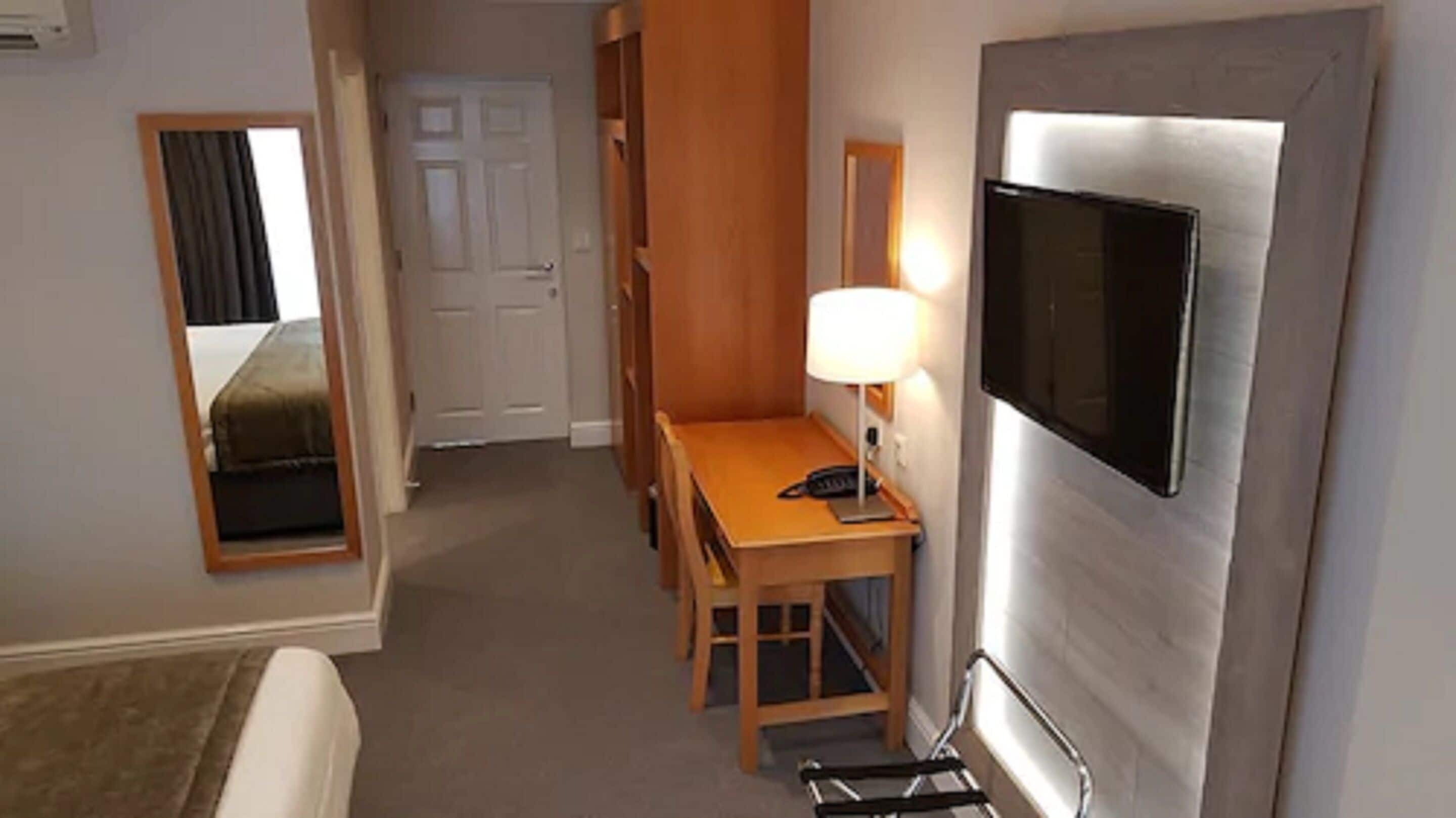 A hotel room with a bed, a desk with a lamp, a TV mounted on the wall, and a door leading to another room.