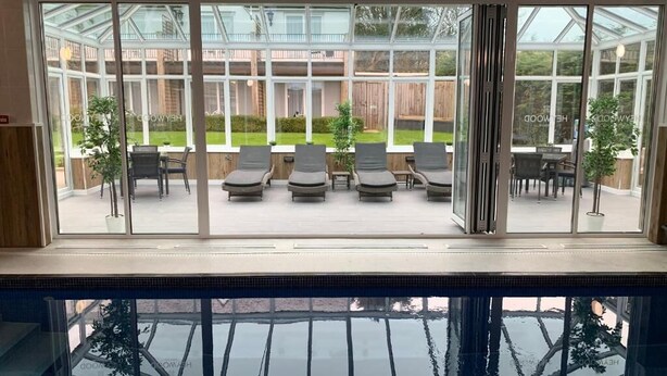 Zona de piscina con cerramiento de vidrio, sillas de descanso y vistas al jardín exterior.