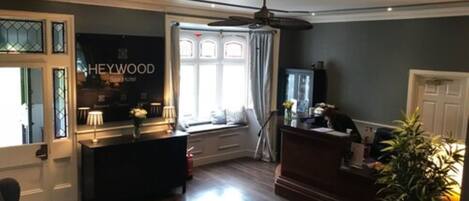 Eine Hotellobby mit einer Rezeption, einer Sitzecke mit Sofa und Sesseln, einem großen Fenster mit Vorhängen und einem Deckenventilator.