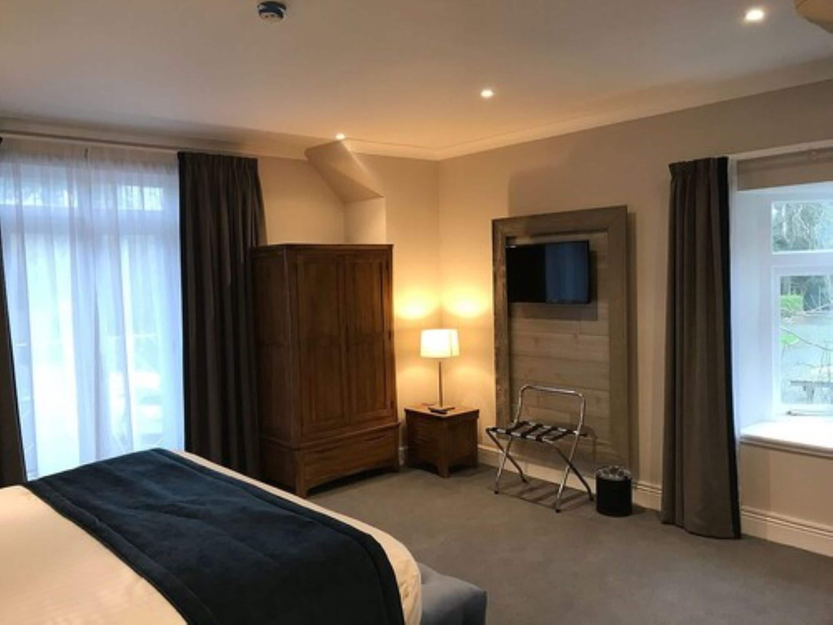 A hotel room with a bed, a wooden wardrobe, a TV mounted on the wall, a small table with a lamp, and a window with curtains.