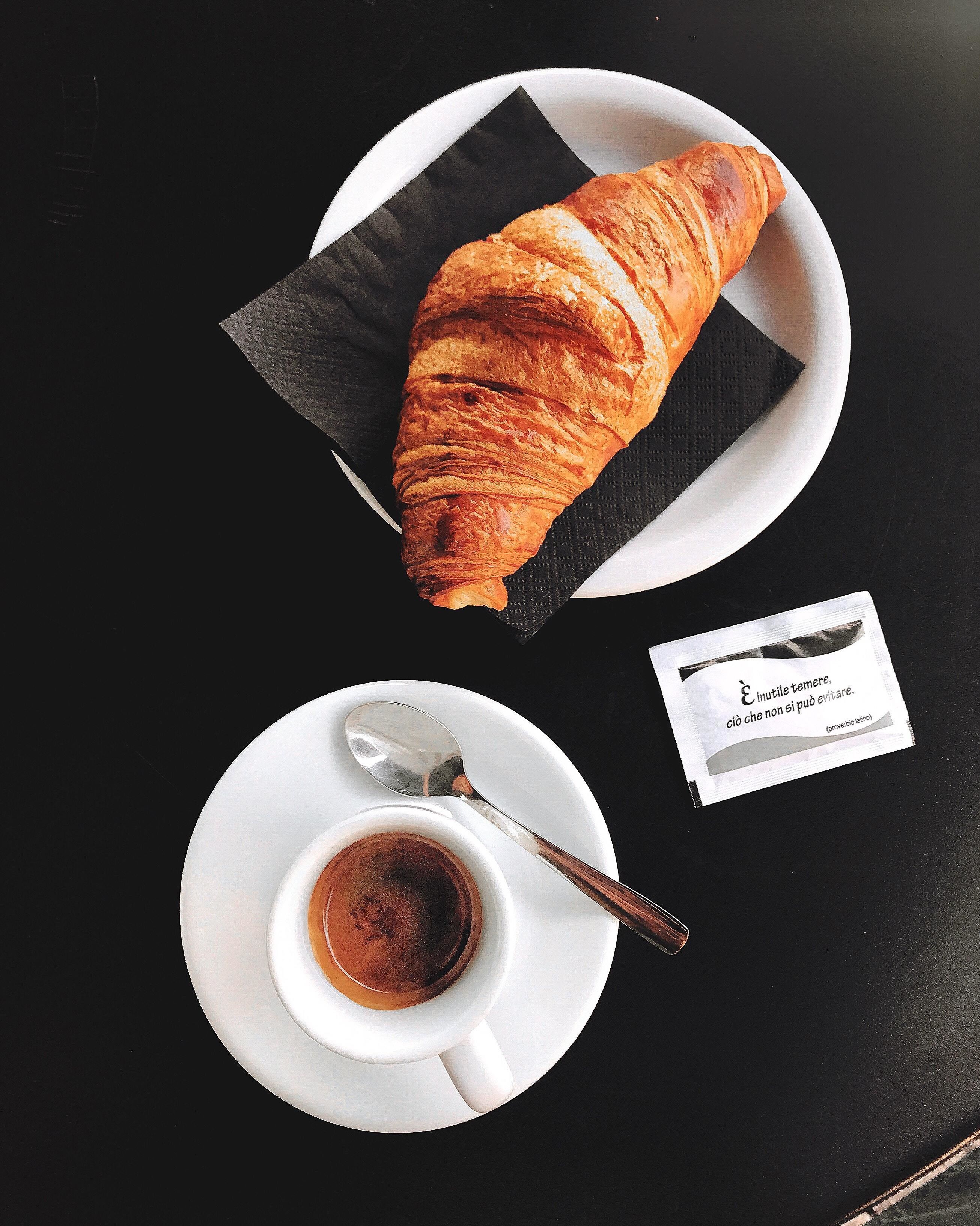 Um croissant em um prato branco, com uma guardanapo, uma colher e uma xícara pequena de café sobre uma mesa preta.