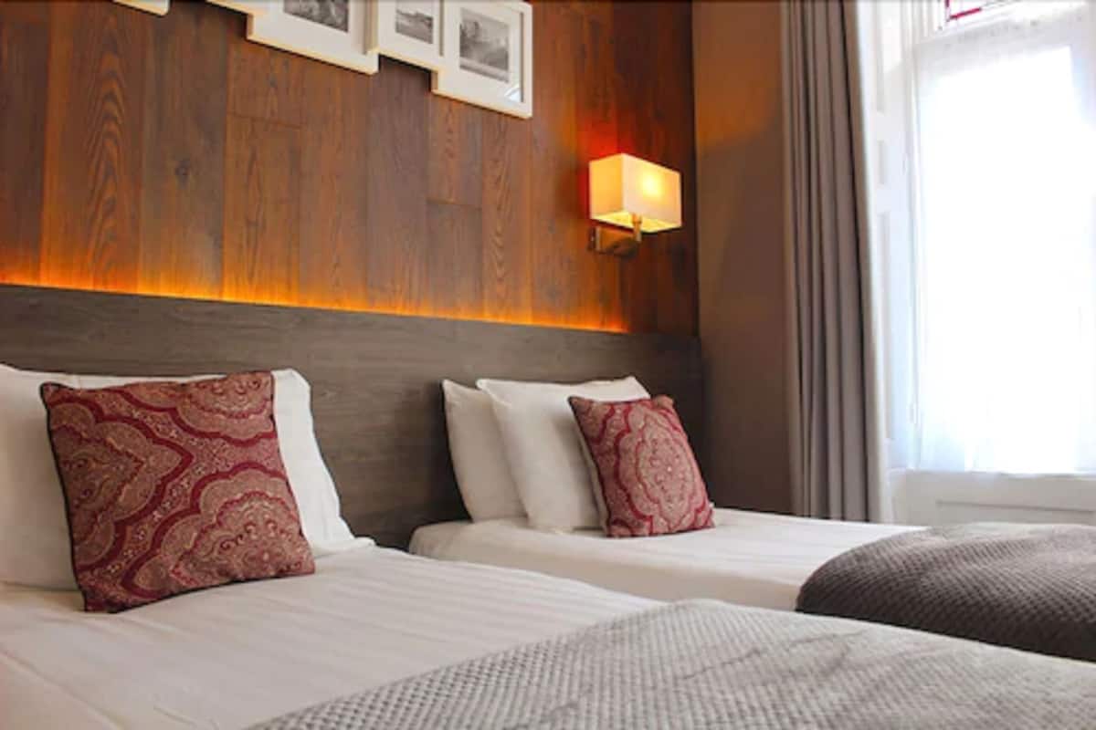 A hotel room with a wooden headboard, two beds with patterned pillows, a wall-mounted lamp, and a window with curtains.