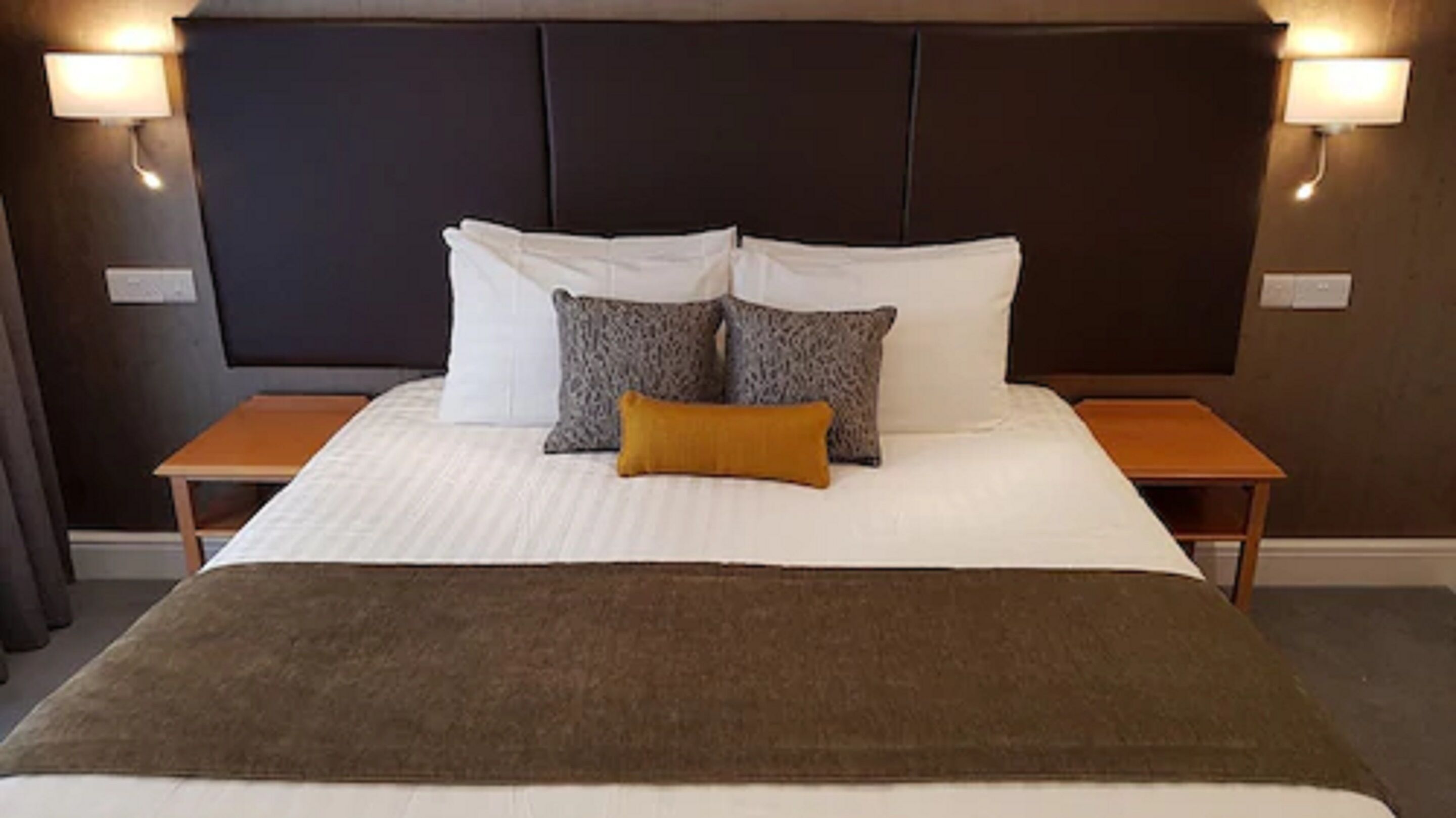 Een net opgemaakt bed met witte en gemêleerde kussens, een bruine plaid en houten nachtkastjes met wandlampen.