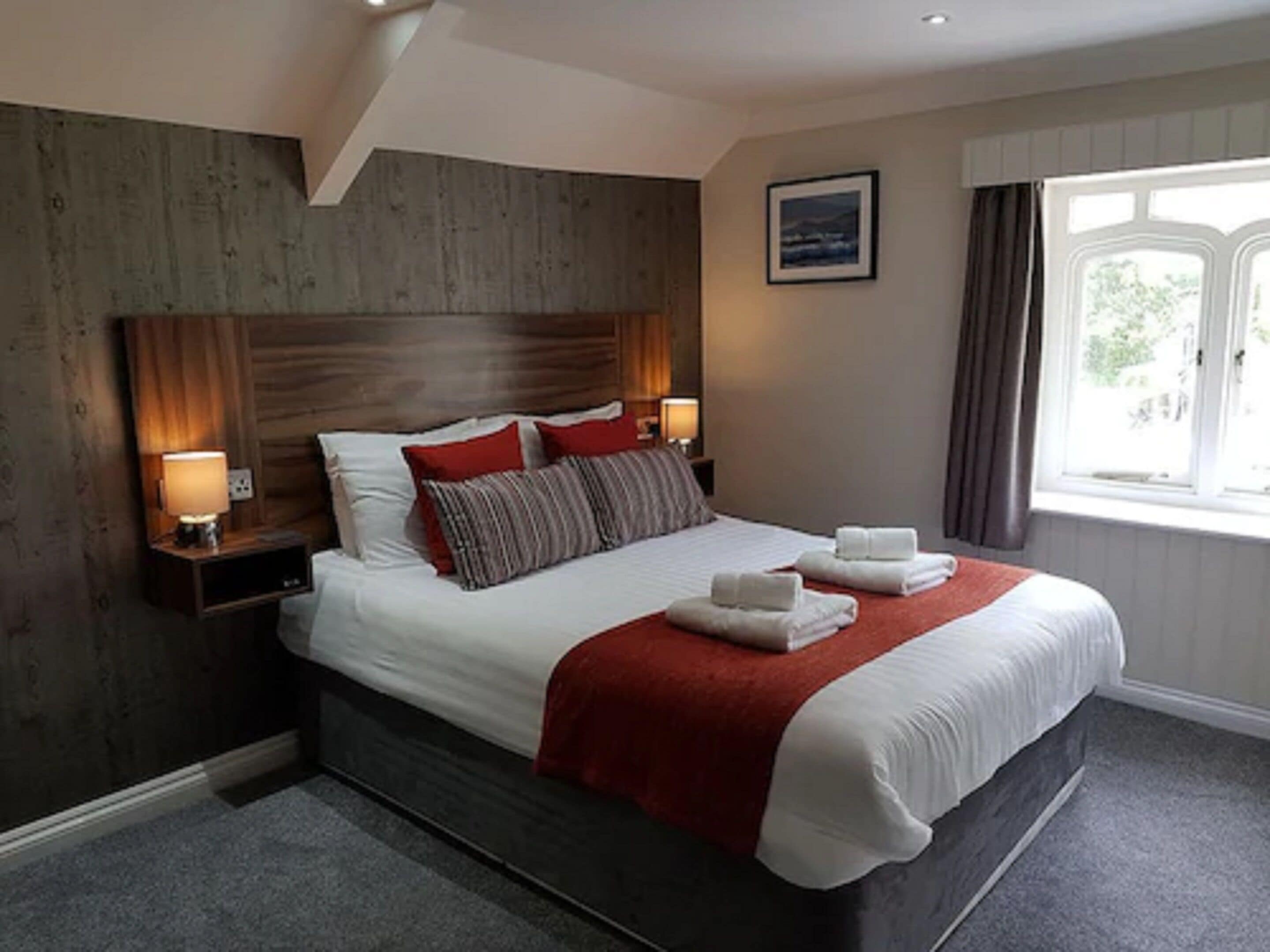 A hotel room with a large bed, wooden headboard, two bedside lamps, a red throw blanket, and a window with curtains.