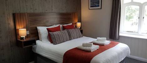 Habitación de hotel con una cama grande, cabecera de madera, dos lámparas de noche, una manta roja y una ventana con cortinas.