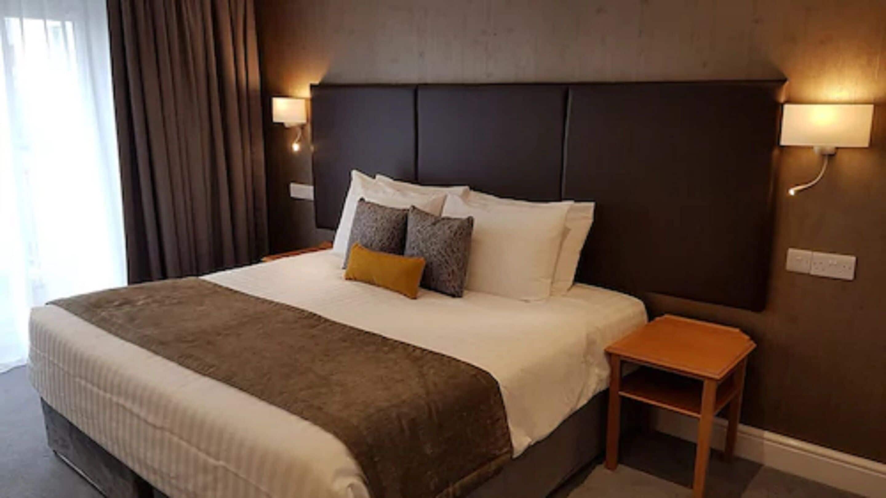 Una habitación de hotel con una cama grande, una mesita de noche de madera y dos lámparas fijadas a la pared.